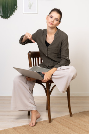 Vista frontale di una giovane donna dispiaciuta seduta su una sedia con un laptop e che mostra il pollice verso il basso
