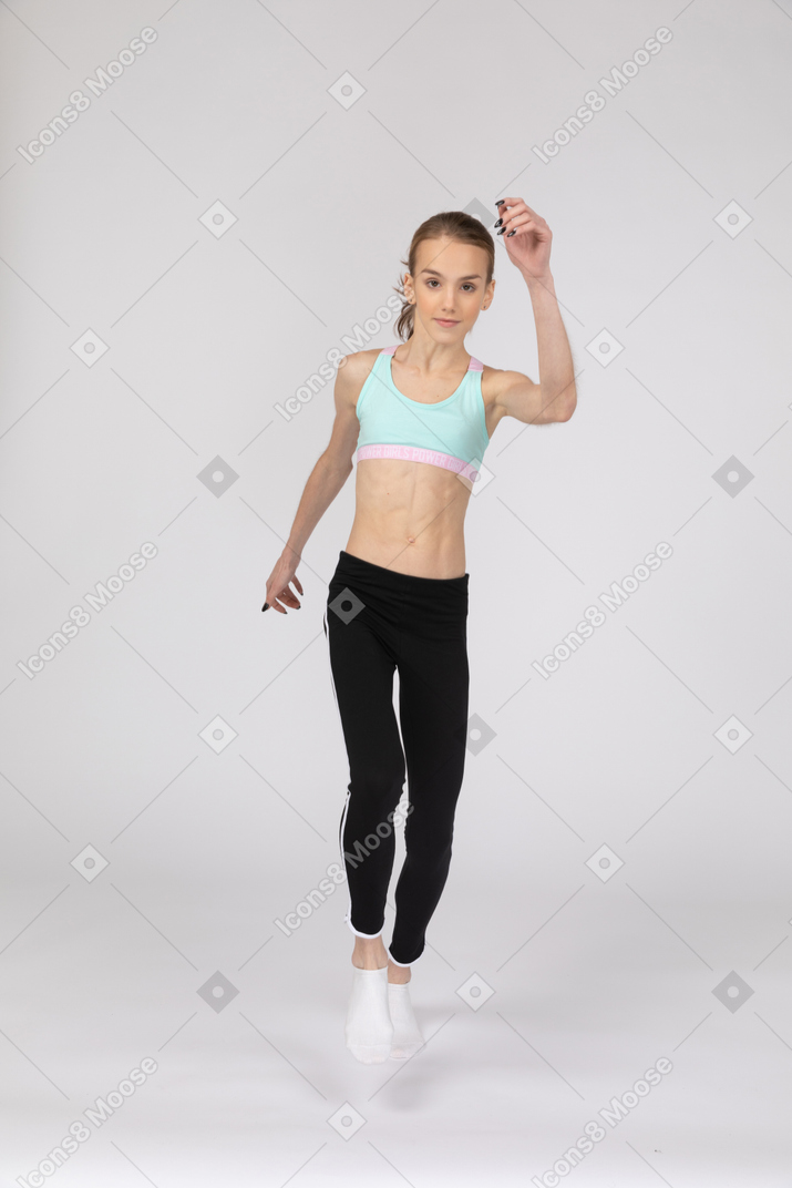 Vista frontal de una jovencita en ropa deportiva levantando la mano y saltando