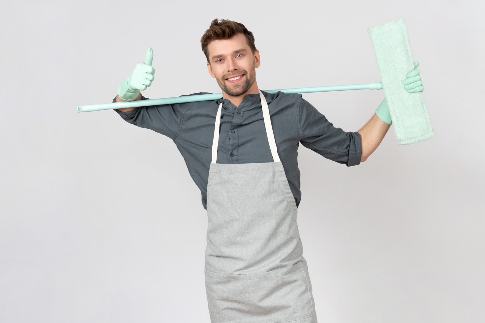 Young househusband holding mop behind his back and sjowing thumbs up