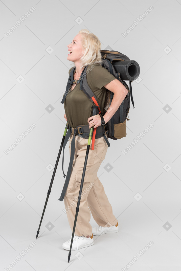 Smiling mature female tourist carrying backpack and walking with walking sticks
