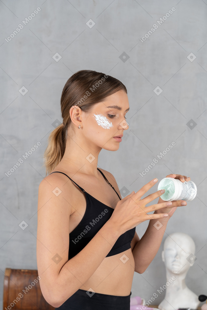 Vista lateral de una joven tomando crema facial del frasco