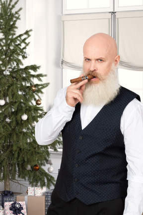 Hombre barbudo serio con un cigarro cerca de un árbol de navidad