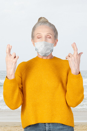 Elderly woman with crossed fingers and face mask standing on a beach