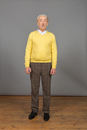 一个po嘴的老男人，穿着黄色套头衫，向前倾斜，看着相机做鬼脸的前视图