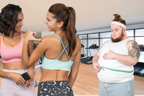 A man measuring his waist and two young women talking to each other in a gym