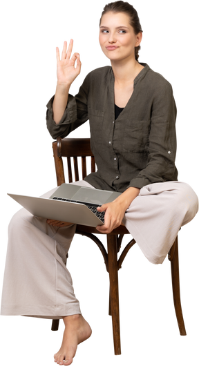 Vista frontale di una giovane donna divertente seduta su una sedia con un laptop e che mostra un gesto ok