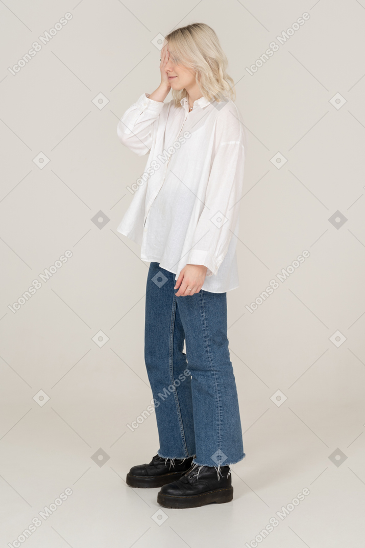 Three-quarter view of a blonde female in casual clothes standing still and hiding face