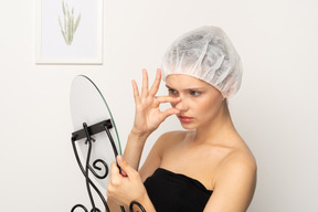 Mujer joven infeliz con gorra médica mirándose en el espejo y tocándose la nariz