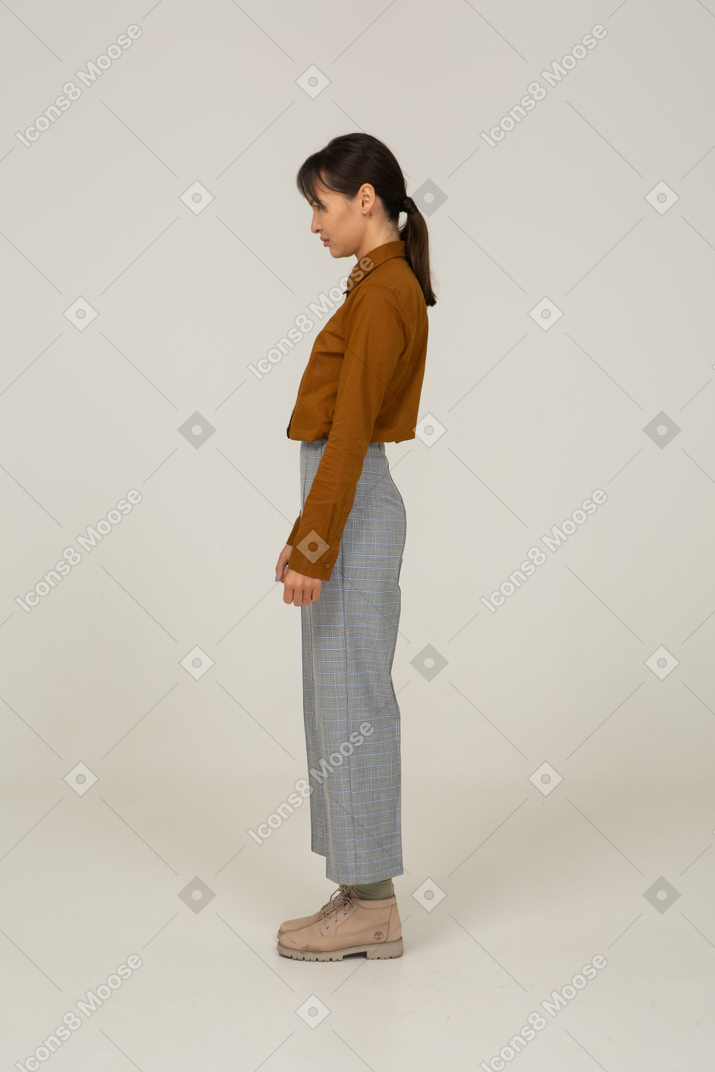 Vista lateral de uma jovem asiática descontente de calça e blusa, parada