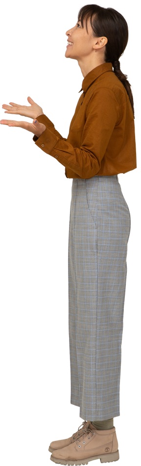 Vista lateral de uma jovem mulher asiática encantada de calça e blusa, levantando as mãos