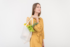 Dreamy young woman holding shopping bag with vegetables and baguette