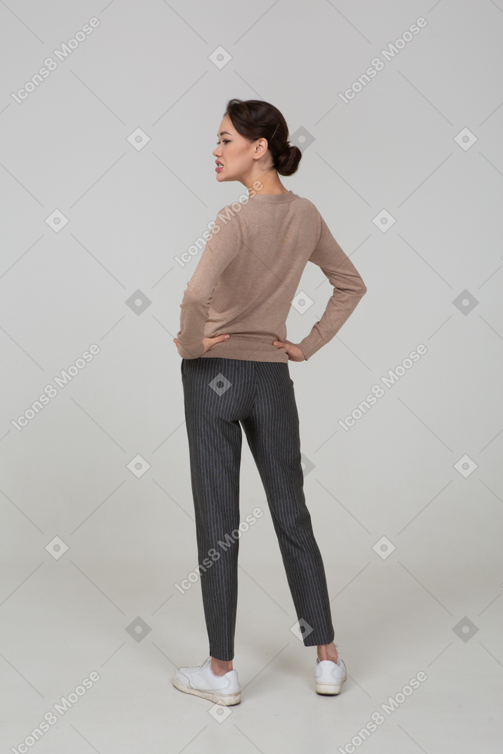 Vista posterior de tres cuartos de una joven en suéter y pantalones poniendo las manos en las caderas y apretando los dientes