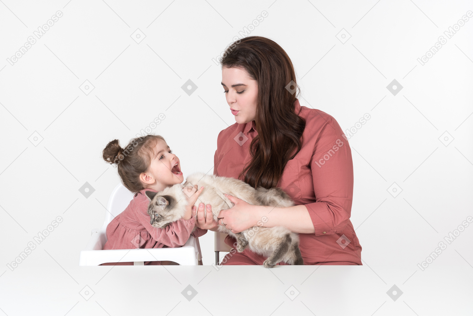 Mother and her little daughter, wearing red and pink clothes, sitting at the dinner table with their family cat