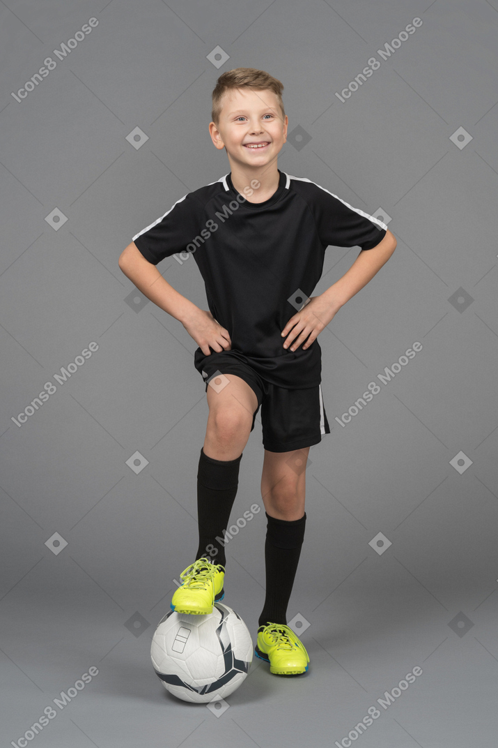 Front view of a smiling child boy in football uniform putting hands on hips and his foot on ball