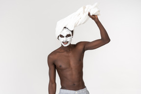 A young black man in grey trousers and a towel on his head going about his skin care routine