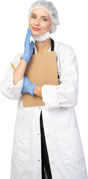 Vista frontal de una joven doctora complacida sosteniendo un lápiz y una tableta y tocando la cara
