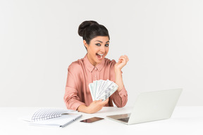 Thrilled young asian woman holding money bills while sitting in front of laptop