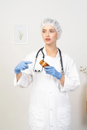 Vista frontal de una joven doctora señalando con el dedo el frasco de pastillas