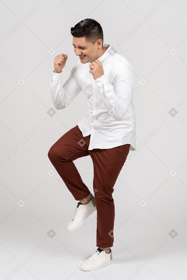 Three-quarter view of a young latino man jumping on one leg and squinting excitedly