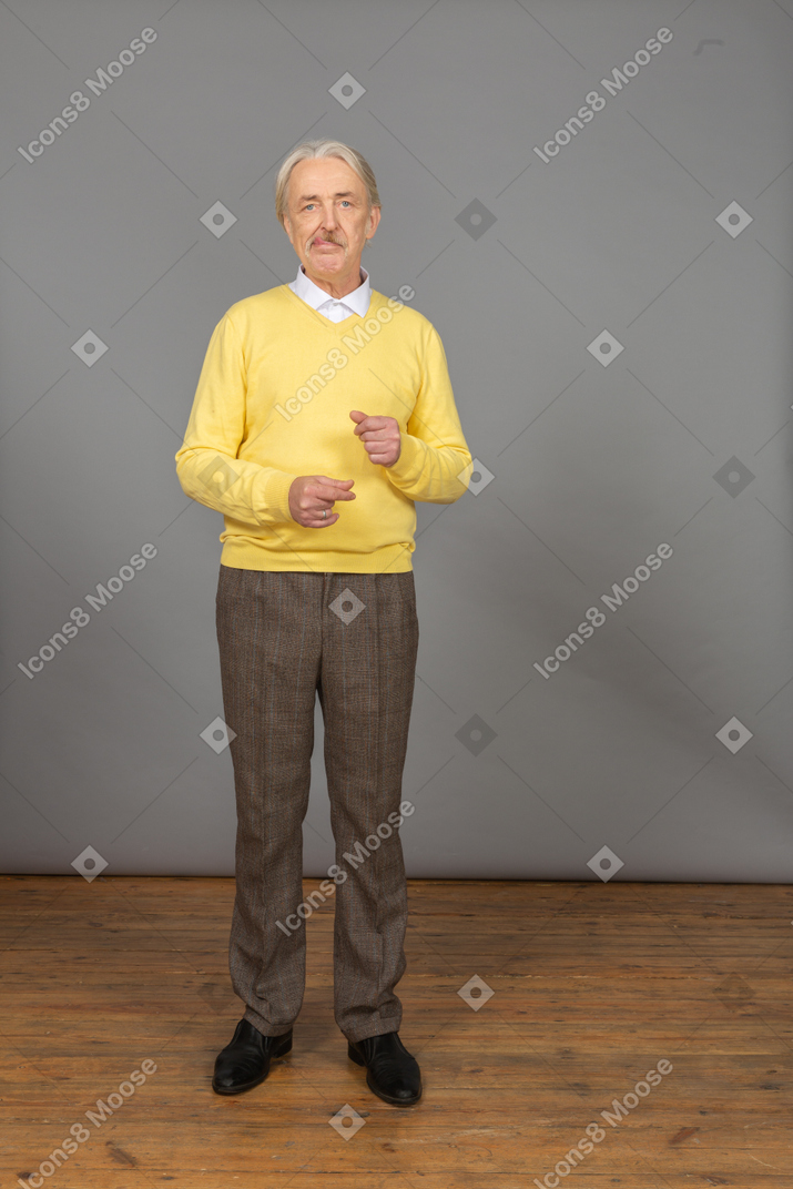 Front view of an old grimacing man in yellow pullover raising hands and looking at camera