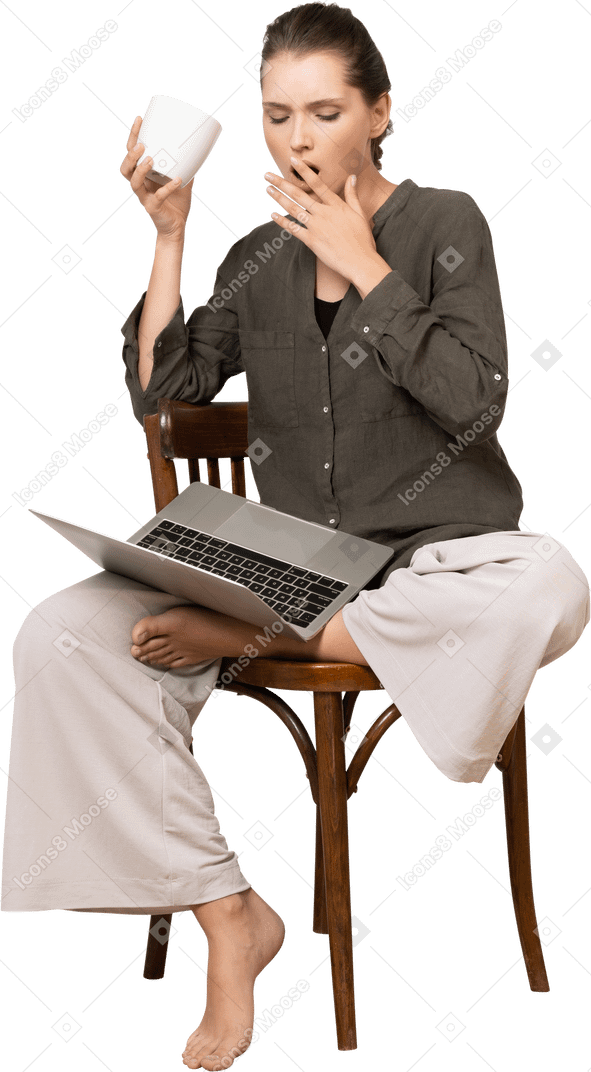 Vista frontal de una mujer joven sorprendida vistiendo ropa de casa sentado en una silla con una computadora portátil y tomando café