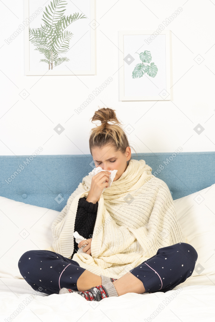 Vista frontal de una joven envuelta en una manta blanca sentada en la cama y sonándose la nariz
