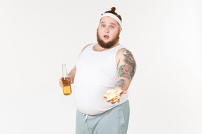 A fat man in sportswear holding a bottle of beer and sharing chips