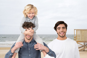 A man with a child on his shoulders next to a smiling man at the beach