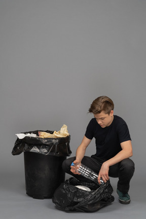 Un jeune homme assis à côté de la poubelle avec une bouteille en plastique dans les mains