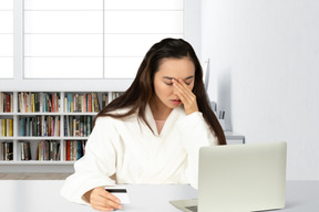 A young woman with facepalm sitting at a desk with laptop holding a credit card