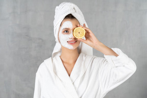 Mujer sonriente en bata de baño sosteniendo un limón sobre su ojo