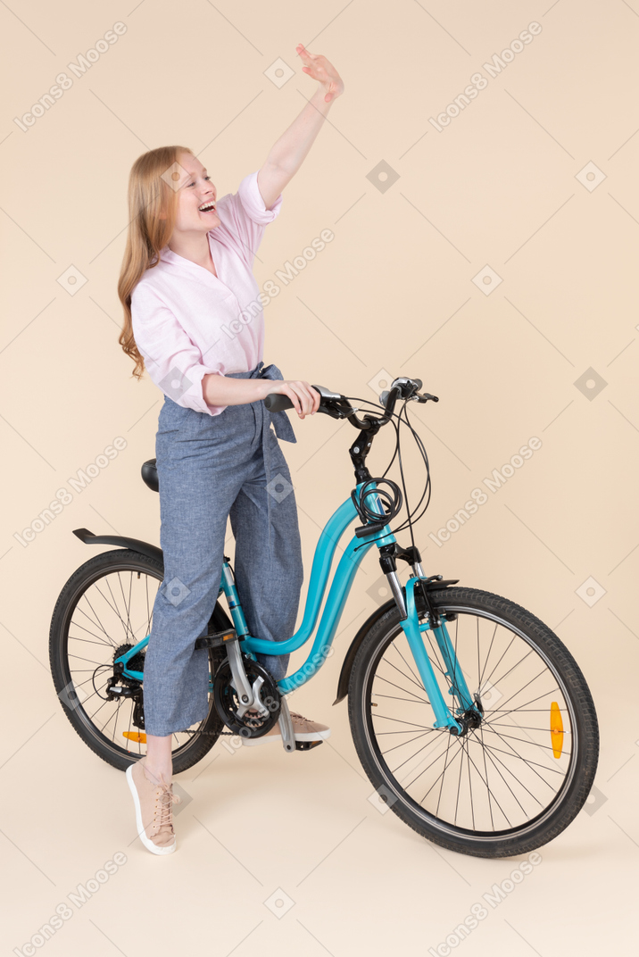 Happy young woman riding on bicycle and waving with a hand