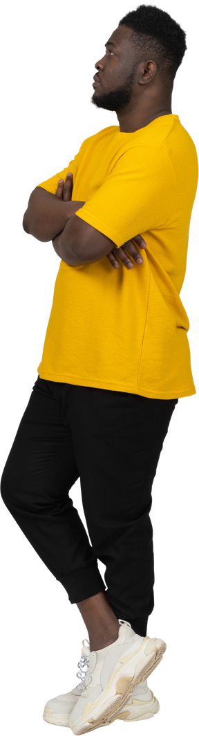 Vista di tre quarti di un giovane uomo sospettoso dalla pelle scura con una maglietta gialla che incrocia le braccia