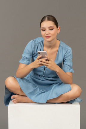 Front view of young woman sitting on a cube and phone messaging