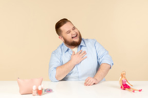 Rindo grande homem sentado à mesa com cosméticos e boneca barbie nele