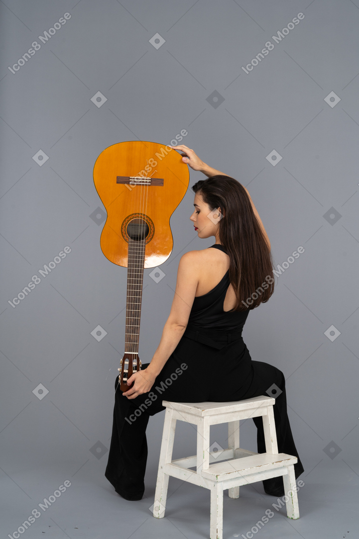 Вид сзади молодой женщины в черном костюме, держащей гитару вверх ногами и сидящей на стуле
