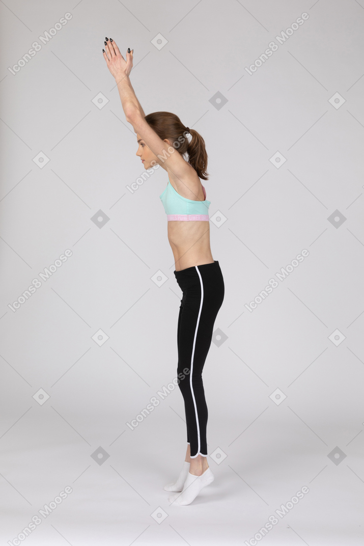Side view of a teen girl in sportswear standing on tiptoes and raising hands