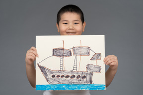 Lächelnder asiatischer junge, der eine zeichnung eines schiffes zeigt