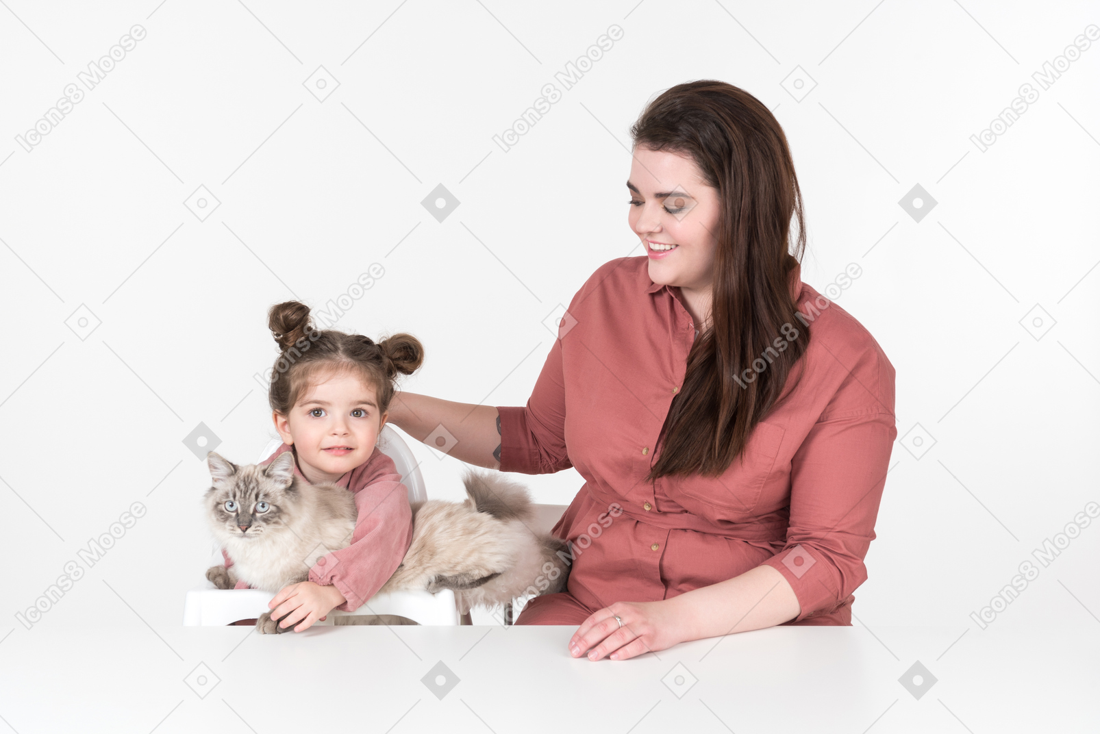Madre y su pequeña hija, vestidas de rojo y rosa, sentadas a la mesa con su gato familiar