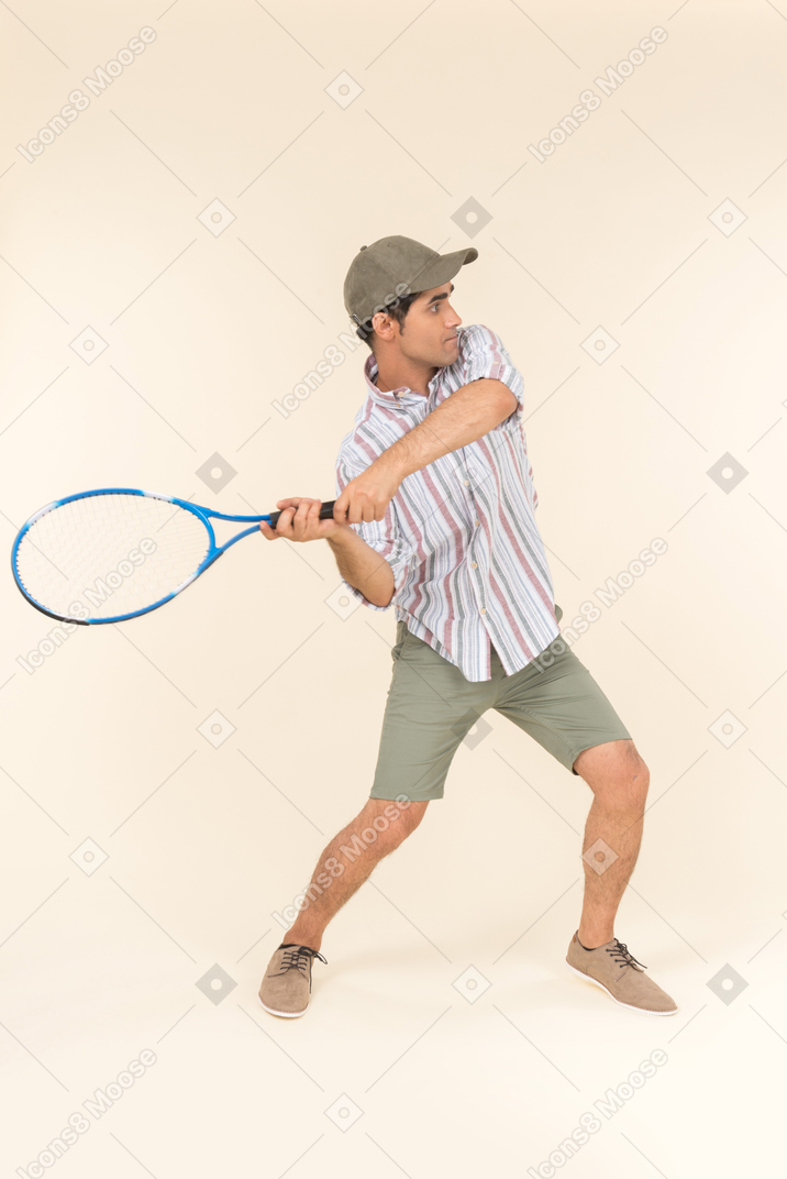 Joven hombre caucásico de pie a medio lado y sosteniendo la raqueta de tenis