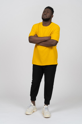 Трехчетвертный вид подозрительного молодого темнокожего мужчины в желтой футболке, скрещивающего руки