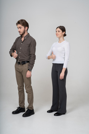 Vista de tres cuartos de una joven pareja alegre en ropa de oficina mostrando el pulgar hacia arriba
