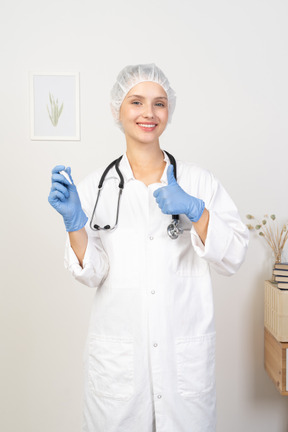 Vista frontal de una sonriente joven doctora con estetoscopio sosteniendo el termómetro y mostrando el pulgar hacia arriba