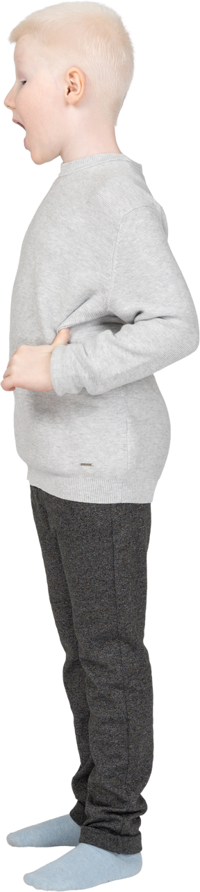 Vista lateral de un niño chico en ropa casual abriendo la boca ampliamente y poniendo la mano en la cadera