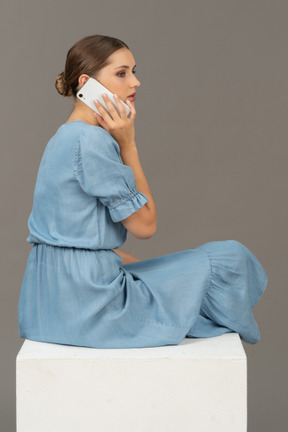 年轻女子坐在立方体上，用智能手机说话的侧背视图