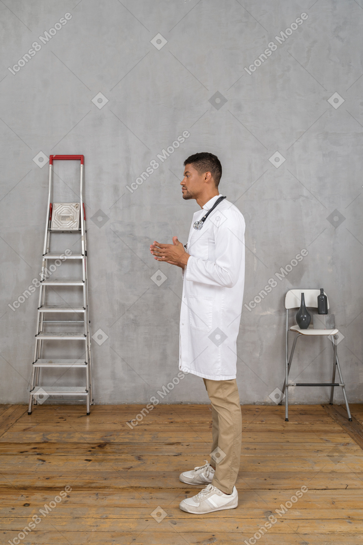 Vista lateral de un joven médico de pie en una habitación con escalera y silla tomados de la mano juntos