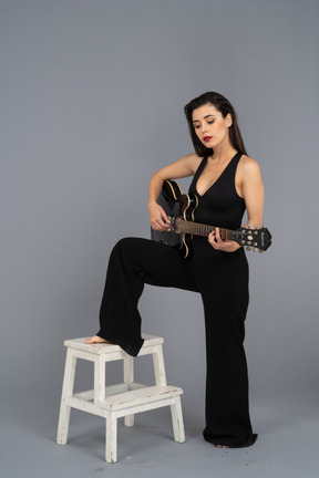 Вид в три четверти молодой женщины в черном костюме, держащей гитару и ставящей ногу на табурет