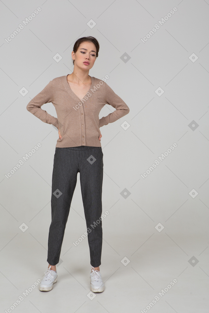 Front view of a tired young lady in pullover and pants putting hands on hips and looking down