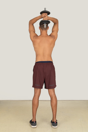 A backside view of the muscular young guy dressed in red shorts with a dumbbell in his hands