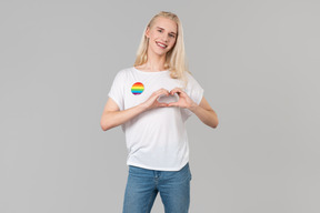 Beau jeune homme aux longs cheveux blonds, debout sur un fond gris, vêtu d'un jean bleu et d'un t-shirt blanc avec écusson lgbt